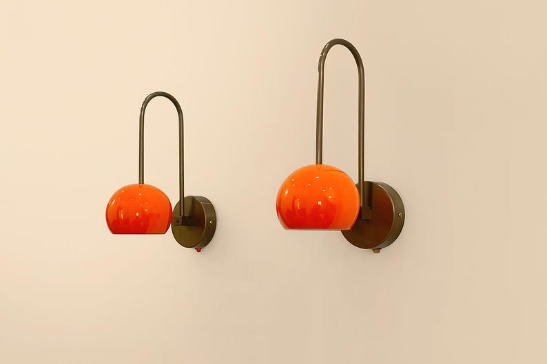May include: Two orange glass globe wall sconces with dark bronze metal accents. Each sconce features a curved arm and a circular base, creating a modern and stylish lighting fixture. The sconces are mounted on a light beige wall.