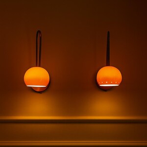 May include: Two orange globe-shaped wall sconces with a dark brown curved arm and a circular base. The sconces are mounted on a warm-toned wall. The sconces emit a warm glow, creating a cozy ambiance.