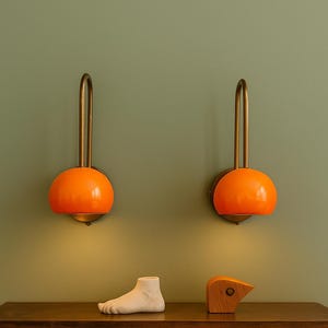 May include: Two orange, mid-century modern wall sconces with brass arms are mounted on a green wall. A wooden shelf below holds a white foot sculpture, a wooden bird sculpture, and a collection of books.