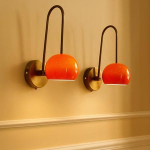 May include: Two modern wall sconces with orange globe shades and dark bronze curved arms. The sconces are mounted on round, brass-colored wall plates. The shades emit a warm glow, creating a stylish and inviting ambiance. The sconces are mounted on a light yellow wall.