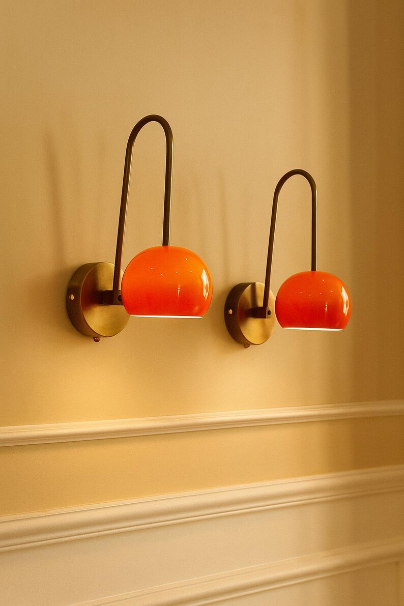 May include: Two modern wall sconces with orange globe shades and dark bronze curved arms. The sconces are mounted on round, brass-colored wall plates. The shades emit a warm glow, creating a stylish and inviting ambiance. The sconces are mounted on a light yellow wall.