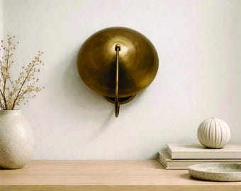 May include: A brass wall sconce with a rounded, disc-shaped shade and a curved handle. The sconce is mounted on a white wall. A vase with dried flowers, books, and a decorative bowl are on a wooden surface below.
