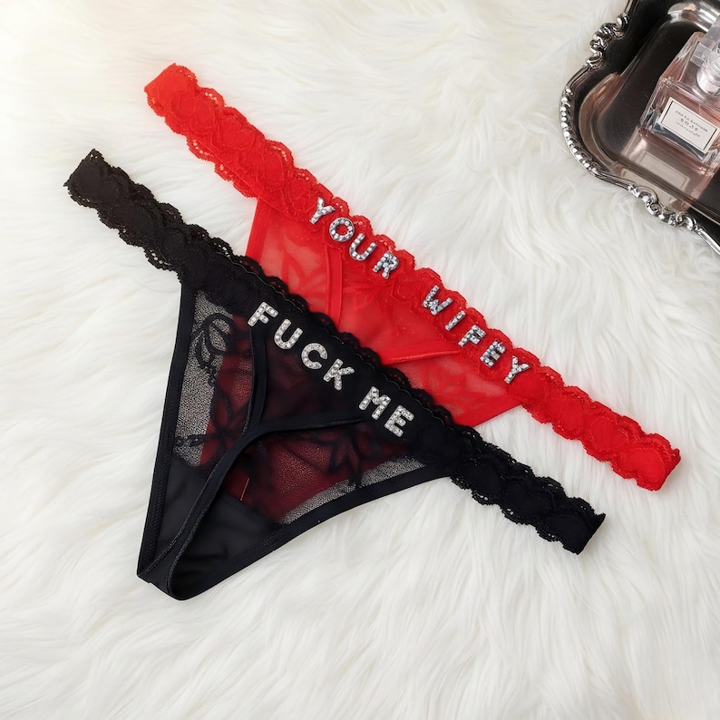 Puede incluir: Dos tangas de lencer&iacute;a, uno negro y otro rojo, con detalles de encaje. El tanga negro tiene el texto "FUCK ME" en pedrer&iacute;a, y el rojo muestra "YOUR WIFEY". Se presentan sobre una superficie blanca y esponjosa.