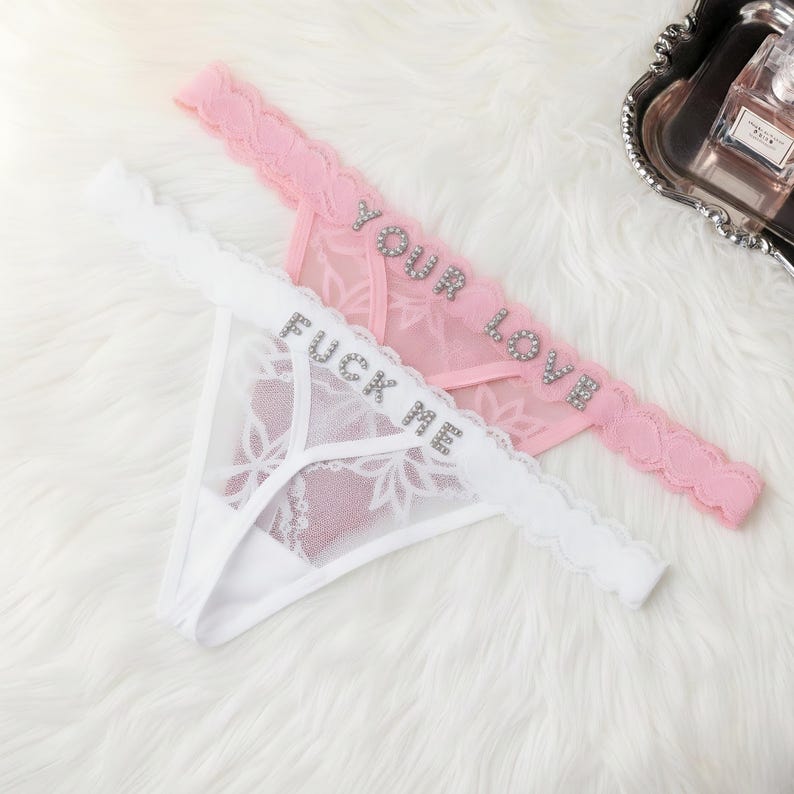 Puede incluir: Dos tangas, uno blanco y otro rosa, con detalles de encaje. El tanga blanco tiene el texto "FUCK ME" en pedrer&iacute;a plateada. El tanga rosa tiene el texto "YOUR LOVE" en pedrer&iacute;a plateada.
