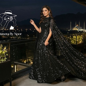 以下が含まれることがあります： 黒のスパンコールイブニングドレス、ケープ付き、ハイネックと流れるようなトレーンが特徴です。 ドレスは夜間に屋外で着用され、街の明かりと背景に建物があります。 ロゴ「La Amora by Noor」が表示されます。