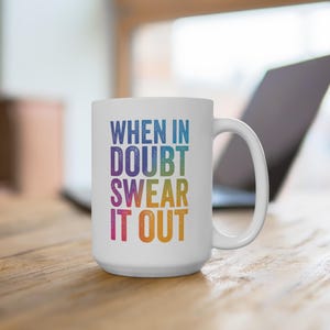 May include: White ceramic mug with the text "WHEN IN DOUBT SWEAR IT OUT" in a rainbow gradient. The mug has a large handle and is sitting on a wooden surface. A laptop is visible in the background.