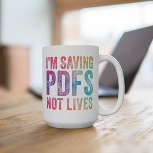 May include: White ceramic mug with the text "I'M SAVING PDFs NOT LIVES" in colorful, distressed lettering. The mug has a large handle and is set on a wooden surface, with a laptop visible in the background.