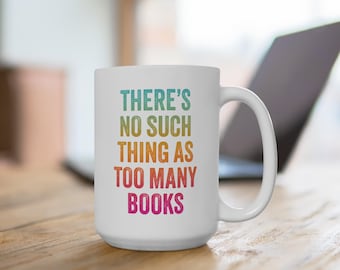 May include: White ceramic mug with the text "THERE'S NO SUCH THING AS TOO MANY BOOKS" in a rainbow gradient. The mug has a large handle and is sitting on a wooden surface.