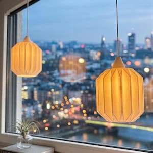 Könnte beinhalten: Zwei cremeweiße Pendelleuchten mit einem gefalteten, geometrischen, hexagonalen Design hängen an dünnen weißen Kabeln. Die Lampen strahlen ein warmes, sanftes Licht aus, das ihre strukturierten, gefalteten Oberflächen vor einer unscharfen Skyline beleuchtet.