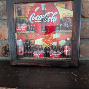 Puede incluir: Un anuncio enmarcado de Coca-Cola con un logotipo rojo y blanco y una ilustración de una botella. La exhibición incluye botellas y cajas de Coca-Cola en miniatura, dispuestas en estantes con detalles de cuerda. El marco es de madera oscura.