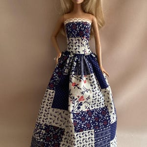 May include: A blonde doll wearing a strapless floor-length gown featuring a patchwork pattern of navy blue and white fabric with small floral and geometric prints, accented by a thin white lace trim along the straight neckline of the bodice.