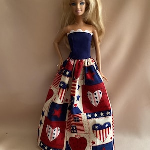 May include: A doll wearing a patriotic-themed dress. The bodice is navy blue with a white scalloped trim, and the skirt features a patchwork design with red, white, and blue stars, hearts, and flag motifs. The doll has blonde hair.