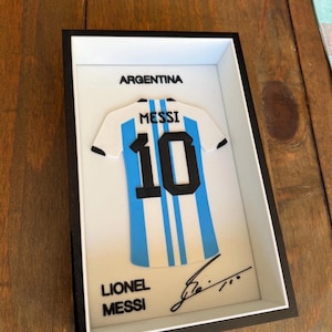Könnte beinhalten: Ein gerahmtes 3D-Wandkunstwerk mit einem weiß-hellblau gestreiften argentinischen Fußballtrikot mit dem Namen MESSI und der Nummer 10 in Schwarz. Der Text ARGENTINA und LIONEL MESSI ist in Schwarz gedruckt, ergänzt durch eine schwarze Tintensignatur.