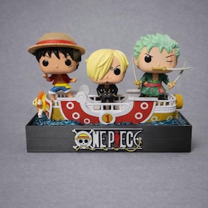 Può includere: Un oggetto da collezione di One Piece con tre personaggi su una nave. I personaggi includono Luffy, Sanji e Zoro. La nave è rossa e bianca con finiture gialle e la base ha il logo One Piece.