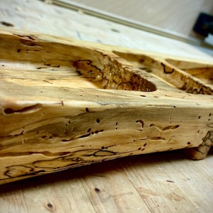 May include: A rectangular wooden tray featuring two carved compartments. The light-colored wood displays a natural grain pattern with dark, irregular streaks and small indentations throughout the surface, showcasing a raw, organic texture.