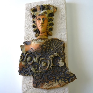 May include: A rectangular wall plaque featuring a sculpted ceramic figure with a tan face and dark, curly hair. The figure wears a textured, dark brown garment with raised circular and geometric patterns, mounted on a rough, off-white base.