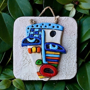 Peut inclure: Sculpture de visage abstrait coloré sur un fond carré texturé. Le visage présente des accents bleus, oranges, rouges et jaunes, avec une base blanche. Une corde marron est attachée pour l'accrocher. L'œuvre est posée sur un fond de feuilles vertes.