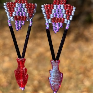 May include: A pair of handmade earrings featuring red, white, and purple beaded starburst designs. The earrings have black V-shaped accents and red or iridescent fish-shaped pendants. The earrings hang from silver-colored hooks.