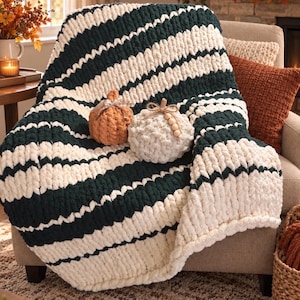 May include: A chunky knit throw blanket featuring thick horizontal stripes in dark forest green and cream. Two decorative fabric pumpkins sit on top of the blanket, one in a muted orange and one in a textured cream with wooden bead accents.