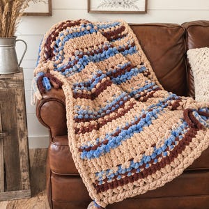 May include: A chunky knit throw blanket featuring horizontal stripes in beige, light blue, and dark brown. The blanket has a textured, looped yarn construction and includes decorative knotted fringe tassels at the corners in matching colors.
