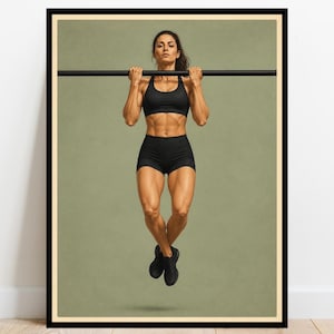 May include: A framed vertical poster featuring a person performing a pull-up on a horizontal black bar. The person wears a black sports bra, black athletic shorts, and black sneakers against a solid muted olive green background.