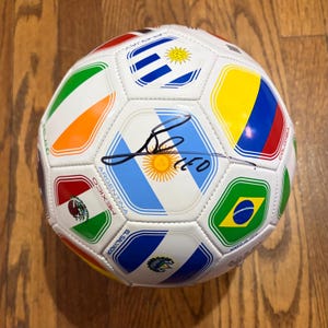Pode incluir: Uma bola de futebol branca com bandeiras de vários países, incluindo Argentina, Brasil e México. A bola tem uma assinatura em tinta preta. Está sobre uma superfície de madeira.
