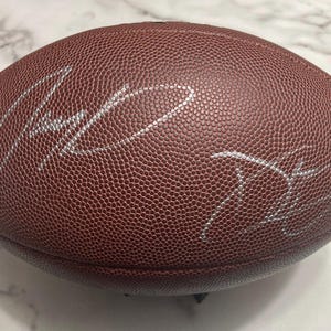 May include: A brown leather football with white signatures. The football has a textured surface and is oval-shaped. The signatures appear to be in silver ink. The football is a sports collectible.