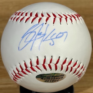 Può includere: Una palla da baseball bianca con cuciture rosse che presenta un autografo a inchiostro blu sulla superficie. Un adesivo olografico circolare nella parte inferiore mostra il testo Total Sports Authentics e il numero 461601, indicando l'autenticazione.