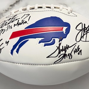 Peut inclure: Un ballon de football américain en cuir blanc arborant le logo bleu et rouge des Buffalo Bills au centre. La surface est couverte de plusieurs signatures et inscriptions à l'encre noire, dont Bills Mafia, HOF 02 et HOF 07, avec des lacets blancs.