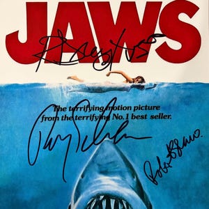 Puede incluir: Un póster de la película Jaws con la icónica ilustración del tiburón y el nadador con el título en letras rojas grandes. El póster incluye los nombres impresos Roy Scheider, Robert Shaw y Richard Dreyfuss, firmado por los tres actores con tinta negra.