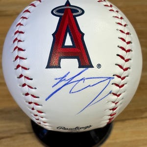 Può includere: Una palla da baseball bianca con cuciture rosse che presenta il logo dei Los Angeles Angels in rosso e blu navy. Sotto il logo c'è un autografo a inchiostro blu. La parola Rawlings è stampata in nero sulla base della palla, poggiata su un supporto.