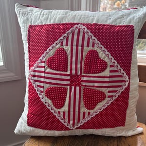 May include: A square pillow with a white background and a red and white patterned design. The center features a red polka dot square with four heart shapes and white stripes. The design is framed with white lace.