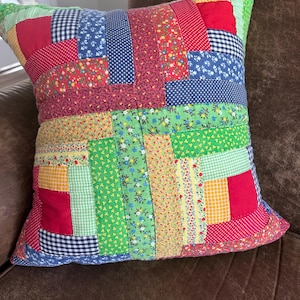 May include: A colourful patchwork cushion with a variety of fabric squares. The cushion features red, blue, green, yellow, and white patterns, including polka dots, floral prints, and gingham. The cushion is square and appears to be handmade.