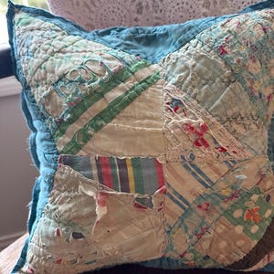 May include: A square patchwork cushion with a teal blue border. The centre features various fabric patches in cream, green, and blue, with red and yellow accents. The fabrics appear aged and distressed, giving a vintage feel.
