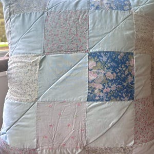 May include: Square patchwork pillow with a variety of floral and solid fabric squares in shades of blue, pink, and white. The pillow has a quilted design with diagonal stitching, creating a textured surface. The pillow is a decorative home accent.