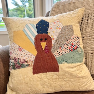 May include: A square cream-colored throw pillow featuring a central turkey appliqué. The turkey body is brown fabric with black button eyes, a yellow beak, and a red wattle. The tail feathers consist of various patterned fabric strips.