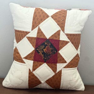 May include: A square quilted throw pillow featuring a central star pattern. The design incorporates cream-colored fabric with leaf-shaped stitching, brown fabric with a fan-like pattern, and a dark center square with a multi-colored leaf print.
