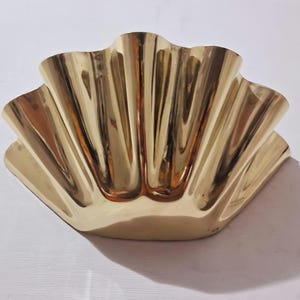May include: A gold-colored, shell-shaped decorative bowl. The bowl has a fluted design with a series of vertical ridges that create a wave-like pattern. The bowl is made of a shiny, reflective material.