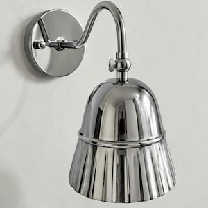 May include: A polished chrome wall sconce featuring a circular mounting plate and a curved arm that supports a bell-shaped shade. The shade has a smooth upper dome and a fluted, pleated lower section, creating a reflective metallic surface.