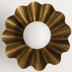 Puede incluir: Aplique de pared de color latón con un diseño ondulado en forma de flor. En el centro, una bombilla esférica blanca emite una luz suave. El aplique está montado en una pared blanca, creando un ambiente cálido.
