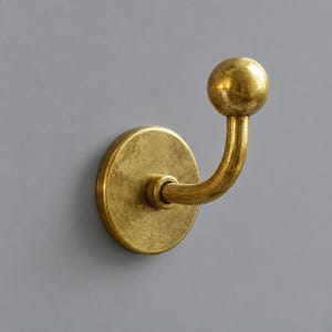 Puede incluir: Un gancho de pared con una placa base circular y un brazo curvo que termina en un pomo esférico. El herraje presenta un acabado metálico en tono dorado antiguo cepillado con una textura visible y una superficie cálida y reflectante.