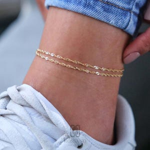 Peut inclure: Un bracelet de cheville doré à double rangée avec un design de chaîne délicate. Le bracelet de cheville est porté sur la cheville, avec une basket blanche et un jean bleu en arrière-plan. C'est un bijou fin.
