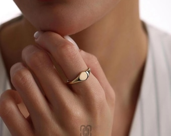 Könnte beinhalten: Ein goldener Siegelring mit glatter, ovaler Oberfläche. Der Ring wird an einem Finger getragen und zeigt seine polierte Oberfläche. Das Design des Rings ist schlicht und elegant, mit einem Band, das sich nach hinten verjüngt. Der Markenname "ROSSANO" ist unten sichtbar.
