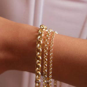 May include: Close-up of a person's arm adorned with four gold chain bracelets. The bracelets vary in style, from thick, rounded links to delicate, thin chains. The warm gold color contrasts with the skin tone and a white background.