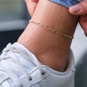 May include: A gold-coloured Figaro chain anklet. The anklet is worn on the ankle above a white trainer. The chain is made of small, interlocking links. The anklet is a delicate piece of jewellery.