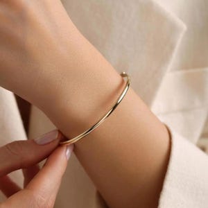 May include: A gold-colored, minimalist bangle bracelet. The thin, curved bracelet is worn on a wrist, with a hand gently touching it. The background is a neutral-toned blazer.