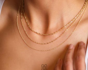 May include: A close-up of a person wearing three delicate gold necklaces. The necklaces have different chain styles, including a rope chain and a simple chain. The necklaces are layered and create a stylish look. The brand name "ROSSANO FINE JEWELRY" is visible.