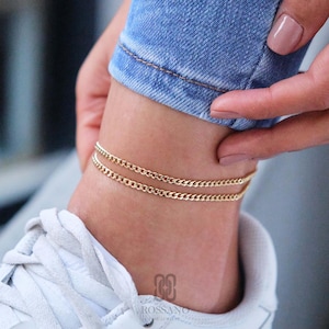 May include: A gold-coloured double-layered chain anklet. The anklet is worn on the ankle, with a white trainer visible below. The person is wearing blue jeans. The brand name "ROSSANO" is visible.