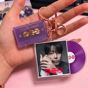 May include: A purple keychain featuring a cassette tape design with the text "YOUR NAME" and a photo of a person. The keychain also includes a purple vinyl record charm and a rose gold clasp.