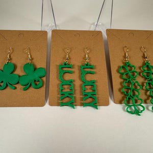 May include: Three pairs of green earrings on display. The earrings include shamrock and "LUCKY" designs. The earrings are displayed on brown cards with gold-colored hooks. The background is white.